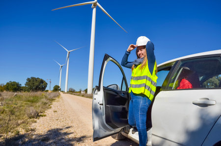 Female engineer adjusting her helmet while exiting a car near wind turbines, symbolizing renewable energy and sustainabilityの写真素材