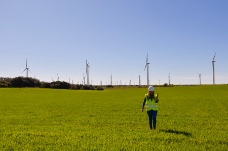 Engineer is walking in a green field with wind turbines generating renewable energy in the...
