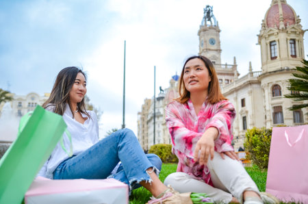 Two happy female friends are enjoying a moment of relaxation on the grass in Valencia city hall square after a shopping spreeの写真素材