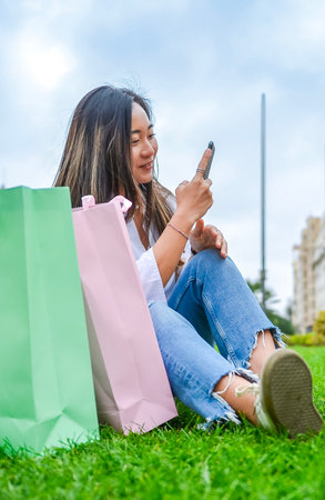 Happy customer browsing on her phone after a successful shopping spree, enjoying a moment of rest in a green parkの写真素材