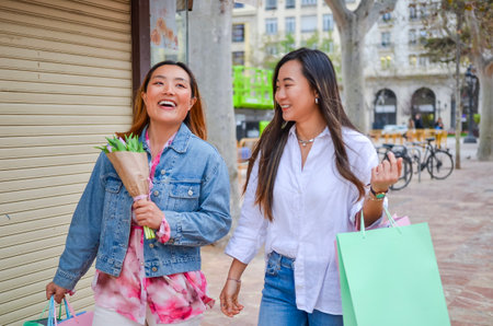 Two cheerful women carrying shopping bags and a bouquet of flowers, enjoying a day of shopping in an urban settingの写真素材