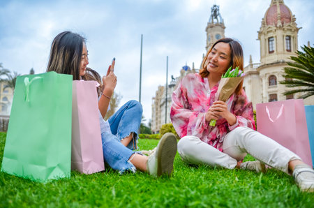 Two female friends are sitting on grass in a park, taking pictures with smartphone and holding shopping bags and flowersの写真素材