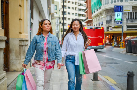Two female friends enjoying a shopping spree, strolling down a city street with colorful bagsの写真素材