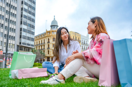 Two asian women relaxing on grass with shopping bags, enjoying conversation after a successful shopping trip in the cityの写真素材