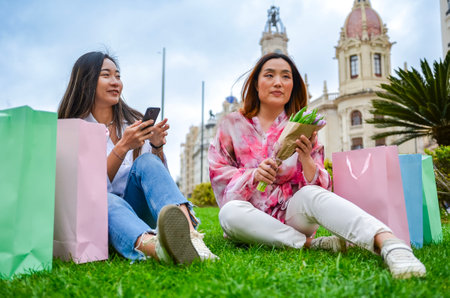 Two happy asian women are sitting on grass with shopping bags, one holding flowers and the other using a smartphoneの写真素材