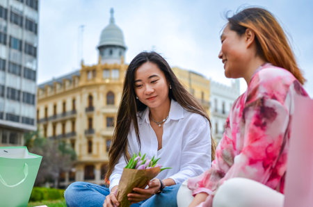 Two female friends sitting on urban park grass, holding shopping bags and a bouquet of flowers, enjoying a relaxing day outの写真素材