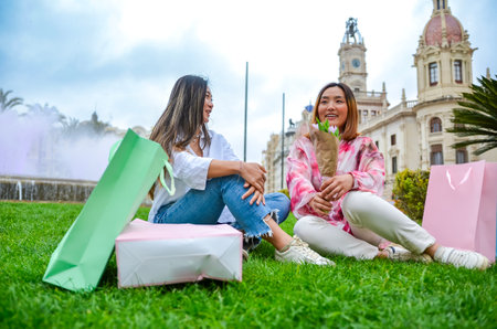 Two young women holding shopping bags and flowers are sitting on the grass in front of the valencia city hallの写真素材