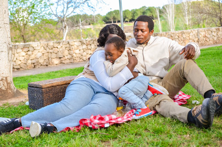 Family enjoying a relaxing picnic in a sunny park, with mother embracing son and father sitting nearbyの写真素材