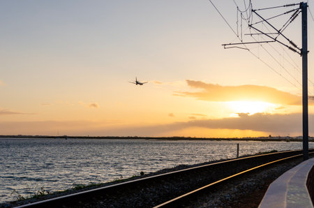 Airplane is descending over a body of water near railroad tracks during a beautiful sunsetの写真素材