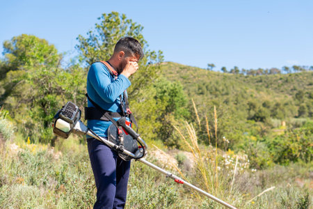 Gardener wiping sweat from his forehead, suffering from heat stroke while using brushcutter in the mountainの写真素材