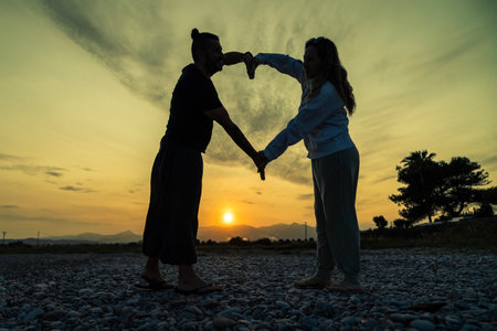 Couple creating heart shape with hands during sunset on a pebbled beachの写真素材