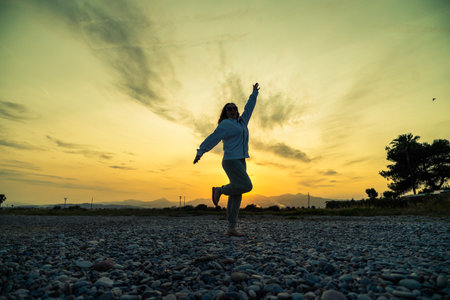 Woman joyfully dancing on a rocky beach during a stunning sunsetの写真素材