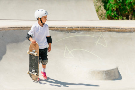 Child skateboarder wearing safety gear holding his skateboard at the edge of a concrete skate park bowlの写真素材