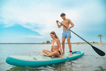 Young man paddling on a sup board with his girlfriend sitting, enjoying a summer day at the beach