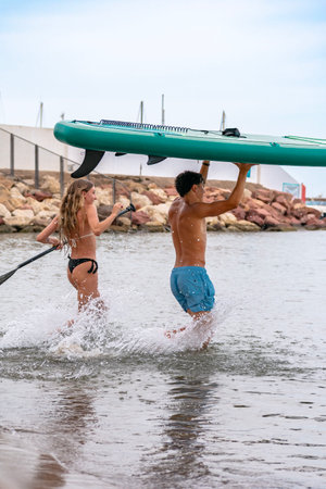 Young couple exiting sea after stand up paddleboarding session, carrying inflatable paddle board and paddleの写真素材