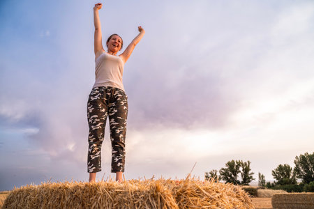 Female farmer standing on hay bale with arms raised, celebrating a successful harvest season at...