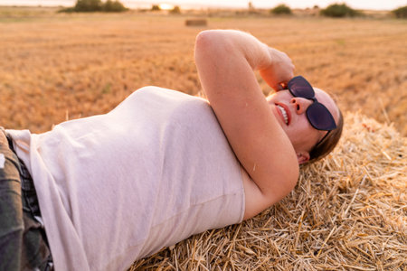 Young woman is lying on a hay bale, enjoying the sunset in her field