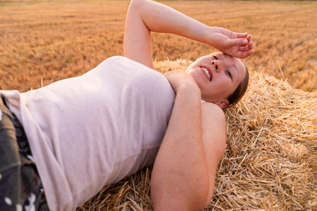Female farmer resting on hay bale, enjoying the golden sunset after a hard day's work in the fieldの写真素材