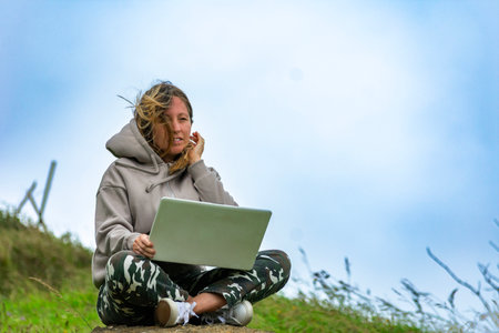 Young woman working remotely using laptop sitting on rock in nature on windy dayの写真素材