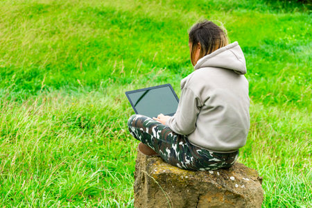 Young woman is working on a laptop while sitting on a rock in a green field, enjoying the freedom of remote workの写真素材