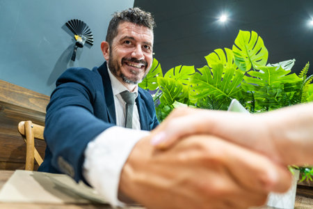 Confident businessman in suit smiles warmly while shaking hands across a deskの写真素材