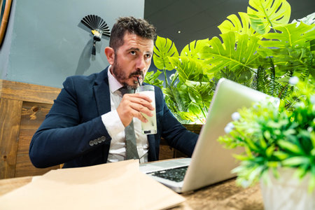 Focused businessman in suit drinking beverage through straw while working on laptopの写真素材