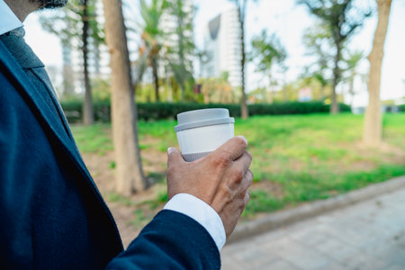 Man in formal suit holding reusable coffee cup while walking through urban parkの写真素材