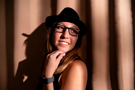 Woman wearing glasses and a hat, hand under chin, standing by a wall with vertical shadows