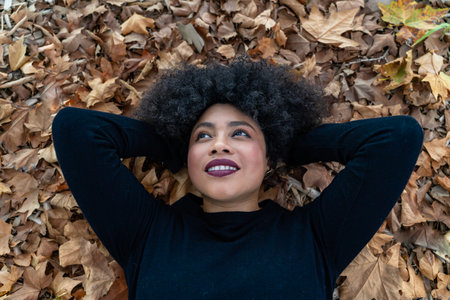 Young black woman relaxing on a bed of dry autumn leaves, looking up with a peaceful smileの写真素材