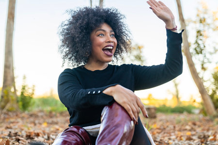 Woman with curly hair waving hand expressing joy and greeting while sitting on autumn leaves