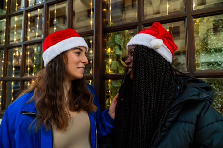 Young friends wearing santa hats talking by festive window lightsの写真素材