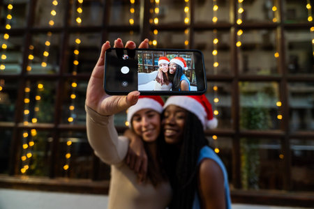 Diverse women wearing santa hats taking a cheerful selfie on a smartphone during christmas celebrationの写真素材