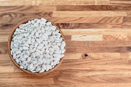 Beans white in a wooden bowl standing on the kitchen table made of oak. Top view.