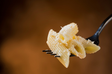 Pasta penne rigate on a fork sprinkled with grated cheese on a beautiful colored background. Close-up.の写真素材