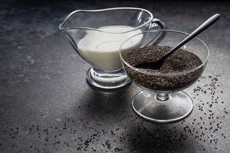 Chia seeds in a glass vase and coconut milk against a dark stone surface with scattered grains.の写真素材