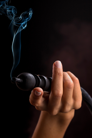 Hookah mouthpiece with a stream of smoke in a beautiful female hand on a dark background. Selective focus.の写真素材