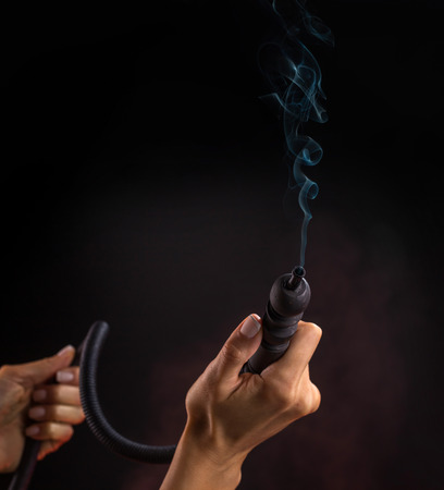 Hookah mouthpiece with a stream of smoke in a beautiful female hand on a dark background. Selective focus.の写真素材