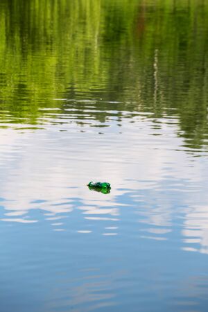 Empty plastic bottle floats on the calm surface of the pond with beautiful reflections of clouds and...