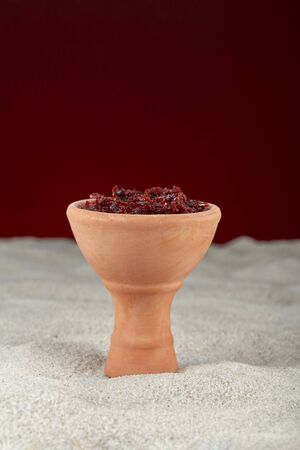 Shisha in a clay bowl, prepared for Smoking hookah, standing on the sand on a maroon background.