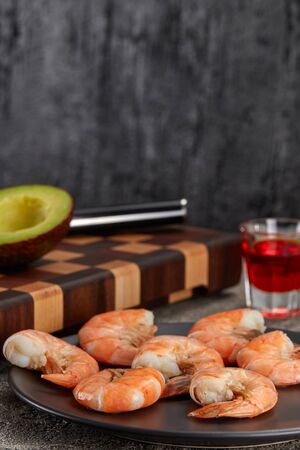 Boiled tails of king prawns on a black plate on the kitchen table against a backdrop of sliced avocado and glasses of drinks.の写真素材