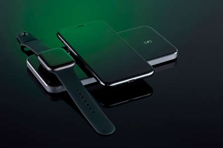 Iwatch with a rubber strap and iPhone X charging on a modern wireless charger on a dark surface with a green glare. Space for your text. Horizontal position. Minsk, Belarus, 1.03.2021.のeditorial素材