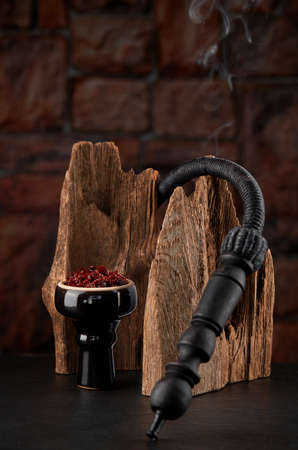 Black glossy hookah bowl filled with apple shisha and prepared for smoking sits on a dark surface next to a textured wood.の写真素材