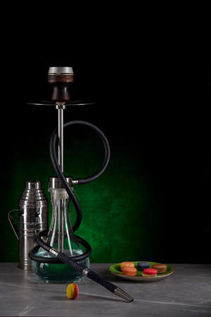 Beautiful modern hookah standing on a table surrounded by a metal protective cap and a plate with colorful macaroons on a dark green background. Space for copying. Vertical location.の写真素材