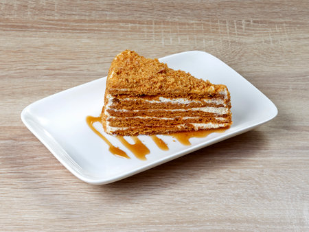Appetizing piece of puff honey cake with cream and sprinkles, on a snow-white rectangular dessert plate standing on a wooden table.の写真素材