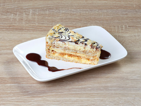 Appetizing piece of puff pastry cake, with cream and chocolate gravy, on a snow-white rectangular dessert plate standing on a wooden table.の写真素材