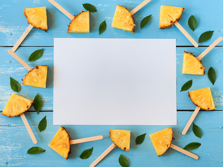 Slice pineapple popsicle sticks and green leafs flat lay on blue wooden table with copy space for summer fruit party concept.の写真素材