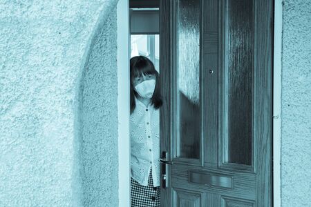 woman in a protective medical mask looking out from behind a partially open door. Coronavirus epidemic UKの写真素材