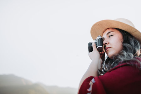 Close up of young latin woman wearing a red poncho and a hat, taking photos with a vintage camera.の写真素材