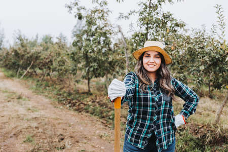 Young smiling latin peasant woman using gardening gloves and leaning on a shovel.