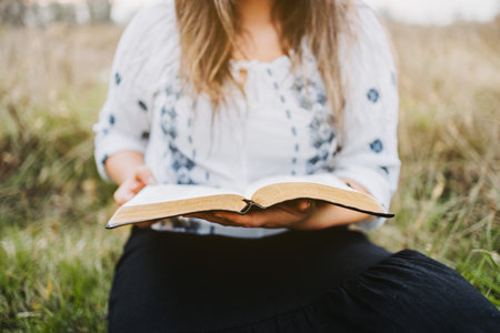 Unrecognizable woman sitting on the grass holding and reading an open Bible. Selective focus.の写真素材